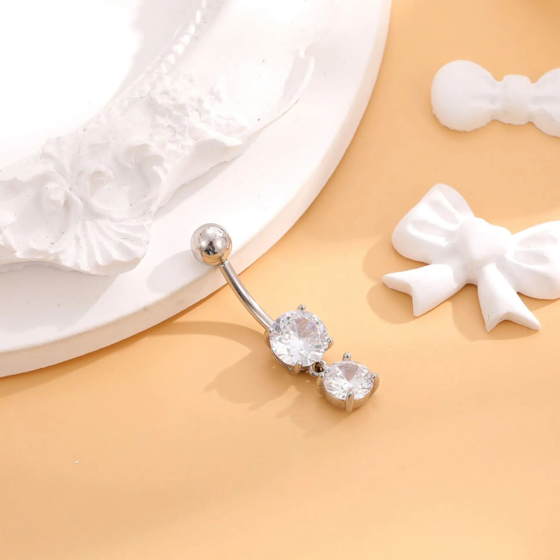Silver belly button ring with clear gemstones on a beige surface with white bows.