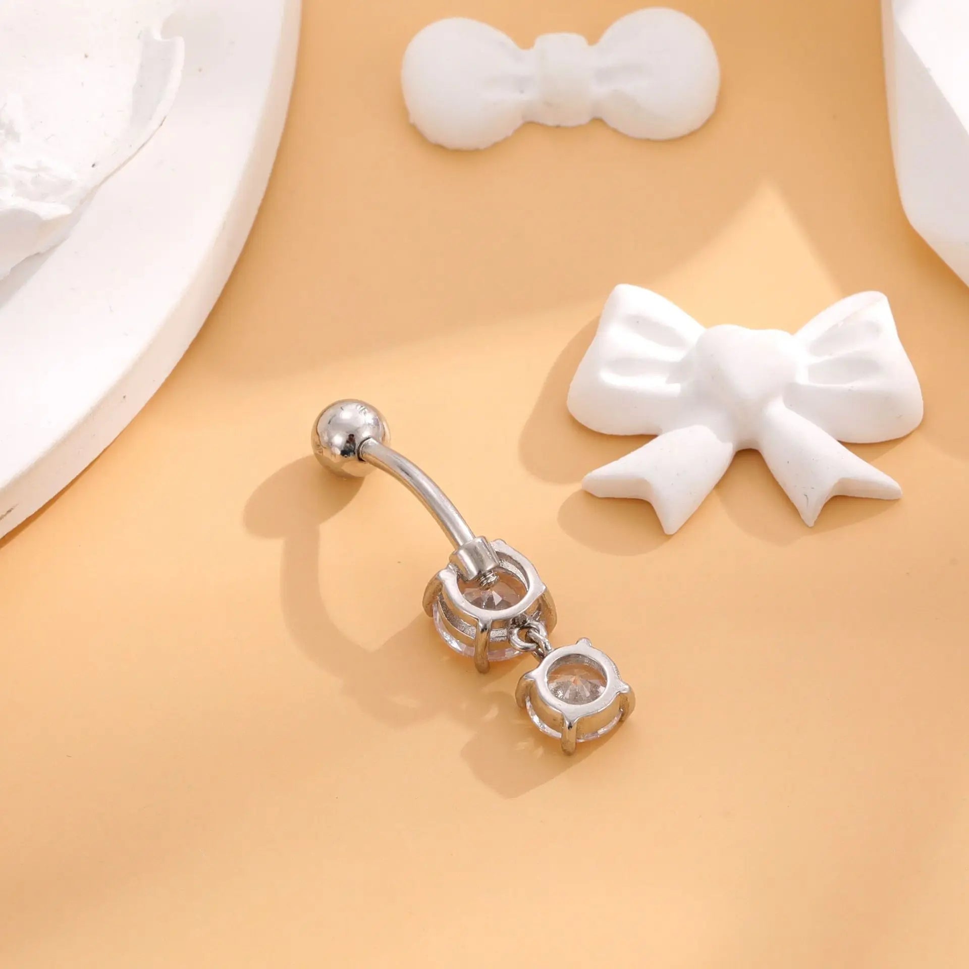 Silver belly button ring with clear gemstones on a beige surface with white bow decorations.