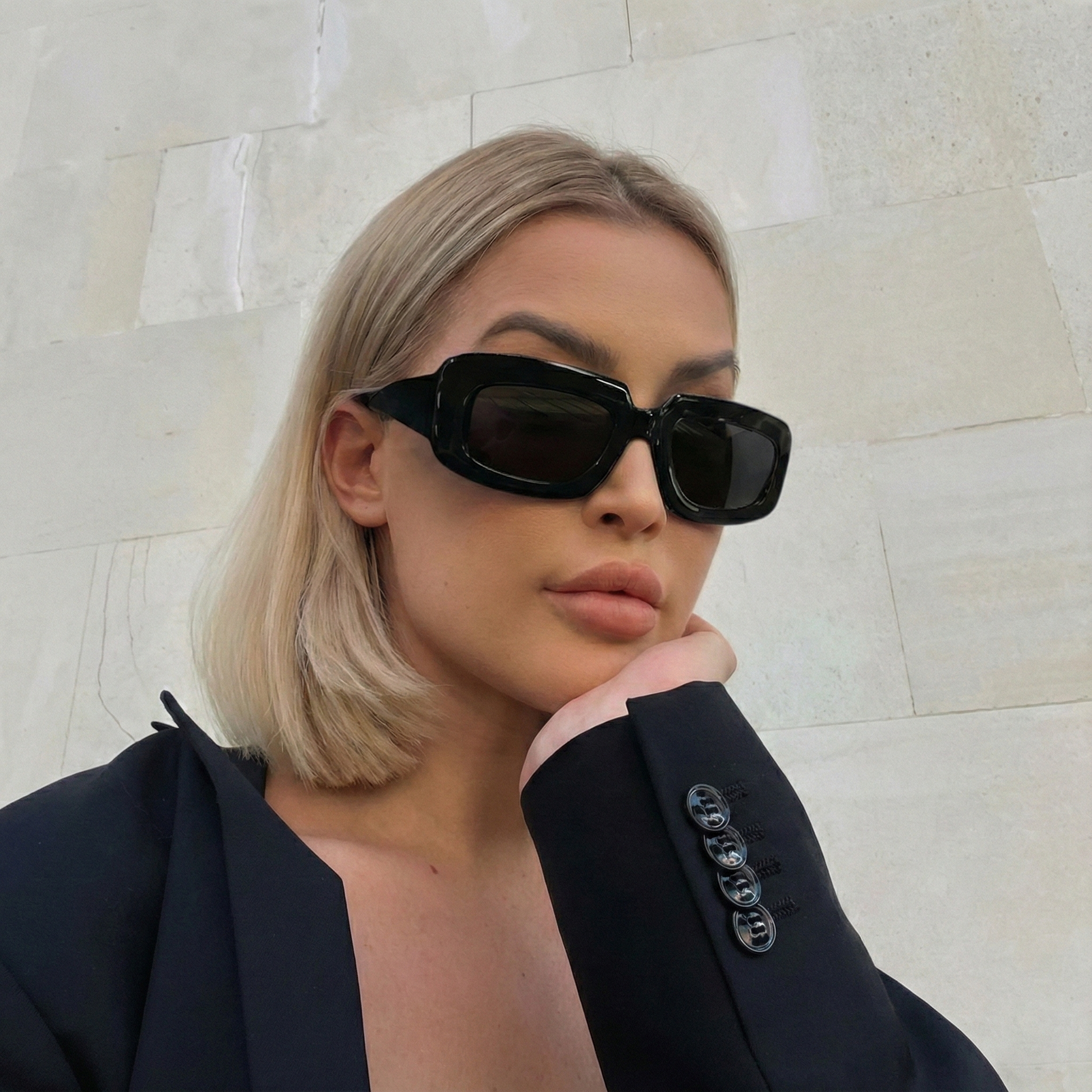 Woman wearing black sunglasses and a dark blazer against a light stone wall.